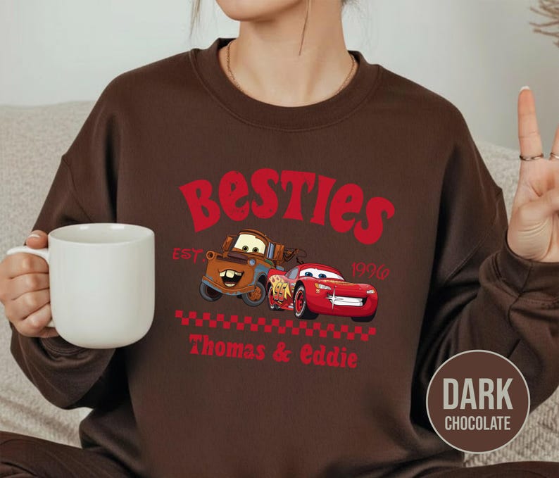 May include: Dark chocolate brown sweatshirt featuring a graphic of two cartoon cars with the text "Besties" in red. The design includes the text "EST 1996" and the names "Thomas & Eddie" in red.