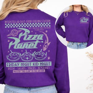 May include: A purple sweatshirt featuring the Pizza Planet logo and alien characters. The back of the sweatshirt has the text "INTERSTELLAR FAST-FOOD FUELING STATION" and "FRIDAY NIGHT KID NIGHT". The front has the Pizza Planet logo.