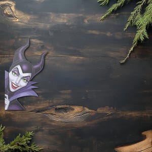 K&ouml;nnte beinhalten: Dekorative Aussparung von Maleficent aus Disneys Dornr&ouml;schen, in Lila und Schwarz mit gr&uuml;nen Augen. Sie befindet sich vor einem dunklen Holzgrund mit gr&uuml;nen Zweigen.