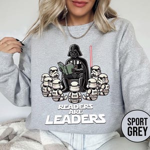 K&ouml;nnte beinhalten: Sportgraues Sweatshirt mit einer Grafik von Darth Vader, der Stormtroopern vorliest. Der Text "Readers are Leaders" steht unter der Grafik. Ein rotes Lichtschwert ist sichtbar. Das Sweatshirt hat lange &Auml;rmel.