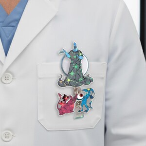 May include: A retractable badge reel with cartoon character designs. The top character is a blue figure in a dark grey robe with green stars. Below are two smaller characters, one red and one blue, with a red eyeball in the middle.