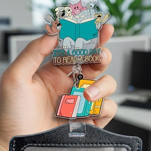 May include: A clear acrylic badge reel featuring a whimsical illustration of various cartoon animals reading a book. The text "IT'S A GOOD DAY TO READ A BOOK" is displayed below the illustration. Attached are colorful book charms.