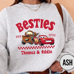 May include: A light gray sweatshirt featuring the word "Besties" in red, with cartoon images of Mater and Lightning McQueen. The text "EST. 1996" and "Thomas & Eddie" are also printed on the sweatshirt.