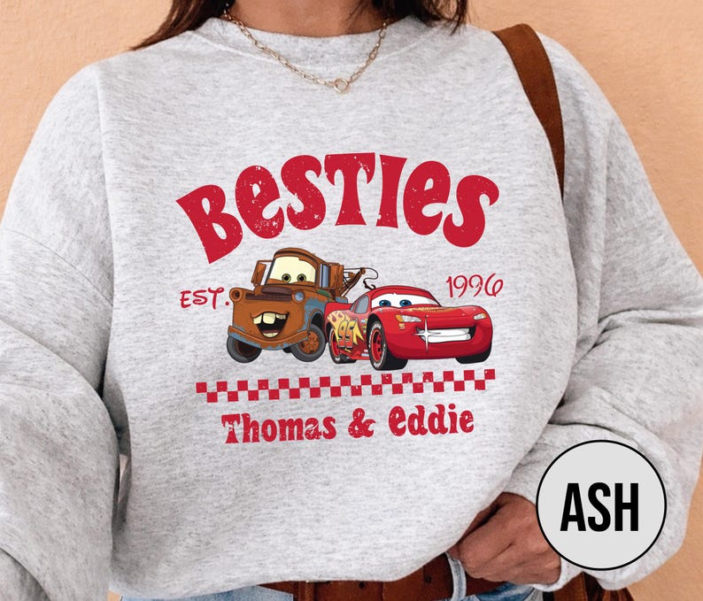 May include: A light gray sweatshirt featuring the word "Besties" in red, with cartoon images of Mater and Lightning McQueen. The text "EST. 1996" and "Thomas & Eddie" are also printed on the sweatshirt.