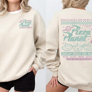 May include: Beige sweatshirt with the Pizza Planet logo on the front and back. The back features the text "INTERSTELLAR FAST-FOOD FUELING STATION" and "FRIDAY NIGHT KID NIGHT" with alien graphics and other text.