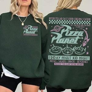 May include: Dark green sweatshirt with the Pizza Planet logo on the front and back. The back features the text "INTERSTELLAR FAST-FOOD FUELING STATION" and "FRIDAY NIGHT KID NIGHT". The design includes alien characters and a space theme.