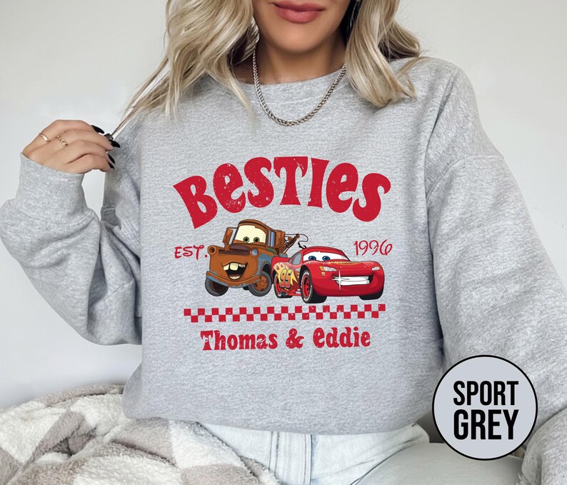 May include: Sport grey sweatshirt featuring a graphic of two cartoon cars with the text "Besties" and "Thomas & Eddie". The design includes the year "1996" and a checkered pattern. The sweatshirt is a crew neck.