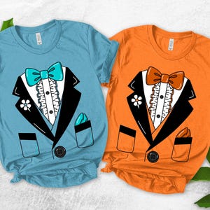 May include: Two short-sleeve t-shirts, one light blue and one orange, each featuring a graphic print of a black tuxedo jacket with a ruffled white shirt front, a colored bow tie, a flower on the lapel, a pocket square, and a single button.