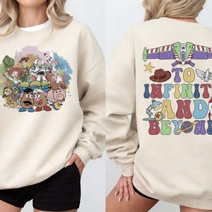 May include: Cream-colored sweatshirt featuring a colorful graphic of Toy Story characters on the front. The back of the sweatshirt has the phrase "TO INFINITY AND BEYOND" with various themed illustrations. A comfortable, casual style.