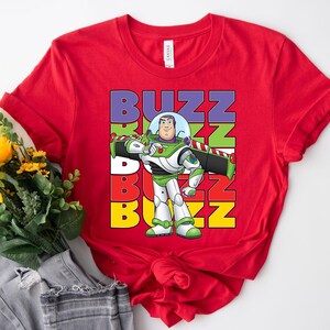 Puede incluir: Una camiseta roja con un gr&aacute;fico de Buzz Lightyear de Toy Story. La palabra "BUZZ" est&aacute; apilada verticalmente en morado, verde, rojo y amarillo. Buzz Lightyear se muestra con su traje espacial verde, blanco y morado.