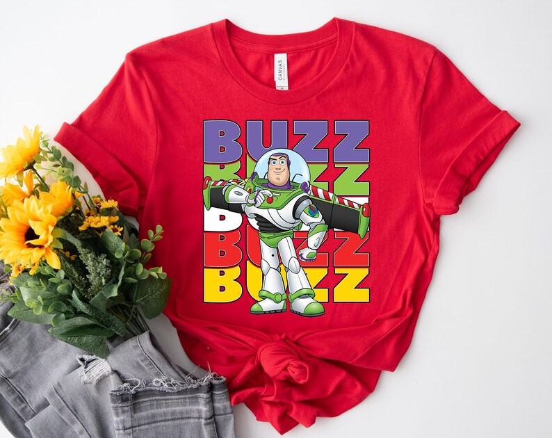 Puede incluir: Una camiseta roja con un gr&aacute;fico de Buzz Lightyear de Toy Story. La palabra "BUZZ" est&aacute; apilada verticalmente en morado, verde, rojo y amarillo. Buzz Lightyear se muestra con su traje espacial verde, blanco y morado.