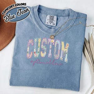 May include: A blue Comfort Colors t-shirt with the word "CUSTOM" in a floral and patterned design. The text "optional line" is embroidered below. A gold necklace rests on the shirt. The shirt is labeled "Blue Jean" in a circular graphic.