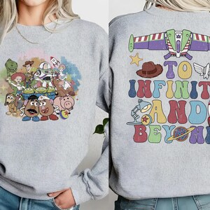 May include: A light gray sweatshirt featuring a colorful graphic of characters from a popular animated film on the front. The back of the sweatshirt has the phrase "TO INFINITY AND BEYOND" with related imagery. The design includes a rocket, a star, and a space ship.