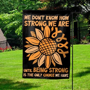 May include: A rectangular garden flag with a black background featuring a large orange sunflower graphic. The text reads WE DON'T KNOW HOW STRONG WE ARE UNTIL BEING STRONG IS THE ONLY CHOICE WE HAVE. An orange awareness ribbon is integrated into the design.