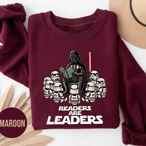 K&ouml;nnte beinhalten: Burgunderfarbenes Sweatshirt mit einer Grafik von Darth Vader, der Stormtroopern vorliest, einem roten Lichtschwert und dem Text "READERS ARE LEADERS" in Wei&szlig;. Das Sweatshirt hat eine einfarbige Burgunderfarbe.