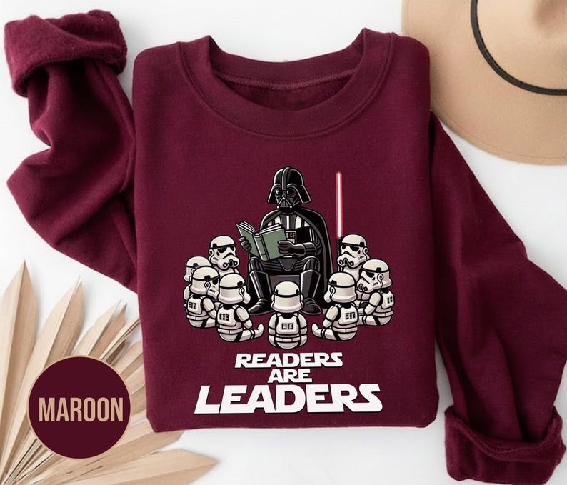 K&ouml;nnte beinhalten: Burgunderfarbenes Sweatshirt mit einer Grafik von Darth Vader, der Stormtroopern vorliest, einem roten Lichtschwert und dem Text "READERS ARE LEADERS" in Wei&szlig;. Das Sweatshirt hat eine einfarbige Burgunderfarbe.