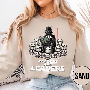 K&ouml;nnte beinhalten: Sandfarbenes Sweatshirt mit einer Grafik von Darth Vader, der Stormtroopern vorliest. Der Text "READERS ARE LEADERS" ist unter der Illustration aufgedruckt. Ein rotes Lichtschwert ist sichtbar.