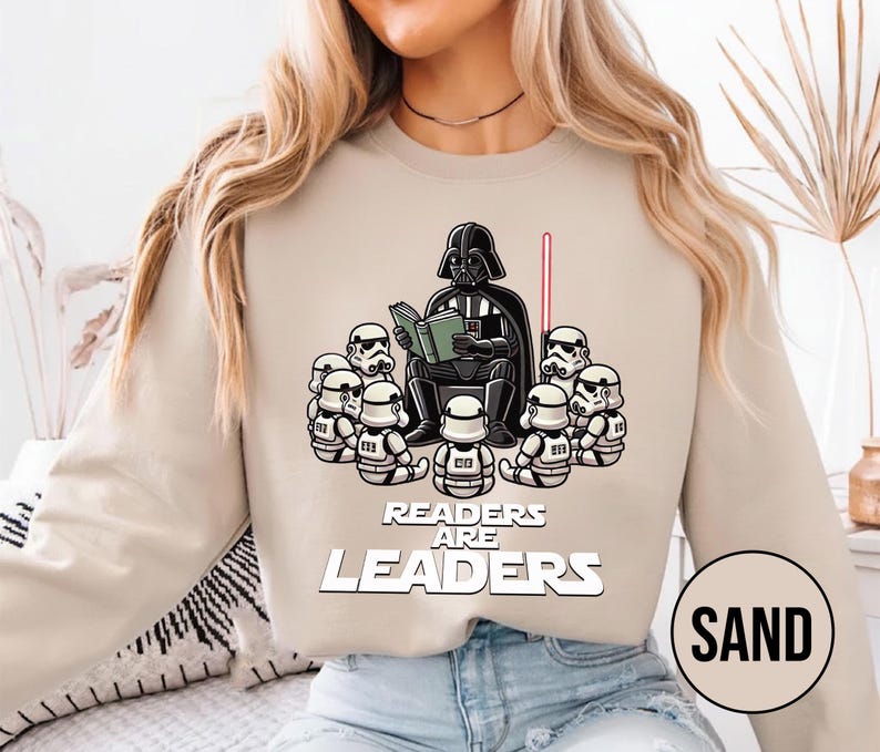 K&ouml;nnte beinhalten: Sandfarbenes Sweatshirt mit einer Grafik von Darth Vader, der Stormtroopern vorliest. Der Text "READERS ARE LEADERS" ist unter der Illustration aufgedruckt. Ein rotes Lichtschwert ist sichtbar.