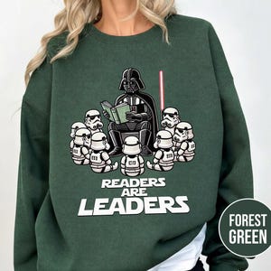 K&ouml;nnte beinhalten: Forstgr&uuml;nes Sweatshirt mit einer Grafik von Darth Vader, der Stormtroopern vorliest. Der Text "Readers Are Leaders" ist in Wei&szlig; gehalten. Ein rotes Lichtschwert ist ebenfalls im Bild zu sehen.