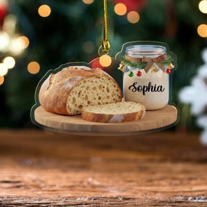 May include: A clear acrylic Christmas ornament featuring a loaf of bread, a slice of bread, and a jar with the name "Sophia." The jar is decorated with a green garland, red and green ornaments, and small gold bells. The ornament is hanging from a gold string.