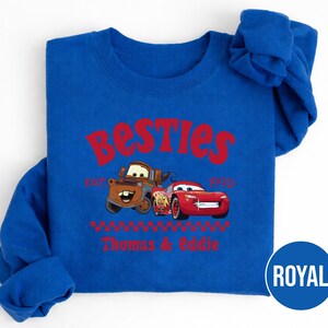 May include: Royal blue sweatshirt featuring the text "Besties" in red, with the phrase "Est. 1910" below. The design includes illustrations of the Cars characters Mater and Lightning McQueen, with the names "Thomas & Eddie" below.