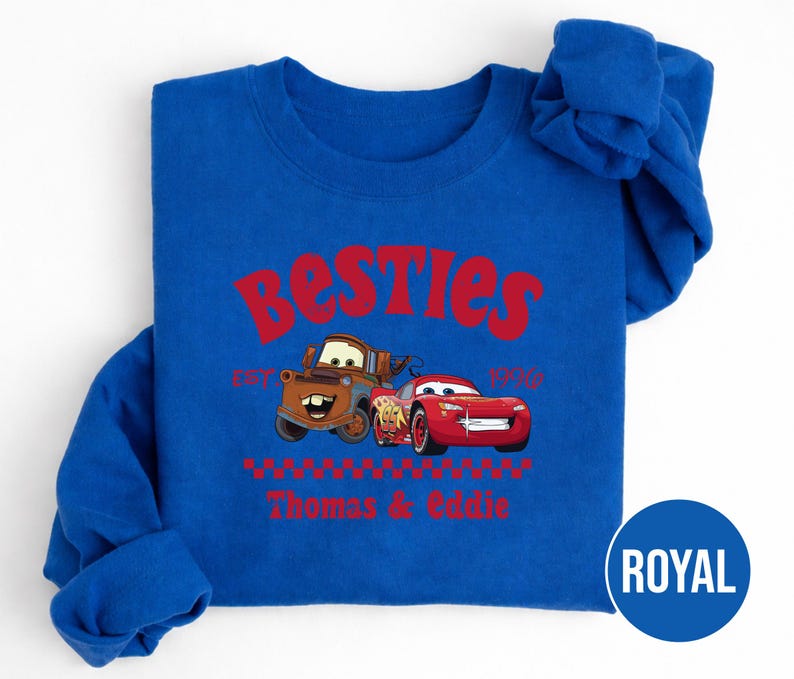 May include: Royal blue sweatshirt featuring the text "Besties" in red, with the phrase "Est. 1910" below. The design includes illustrations of the Cars characters Mater and Lightning McQueen, with the names "Thomas & Eddie" below.