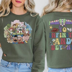 May include: Olive green sweatshirt featuring a colorful graphic of Toy Story characters on the front. The back of the sweatshirt displays Buzz Lightyear's spaceship and the phrase "To Infinity and Beyond" in a retro font.