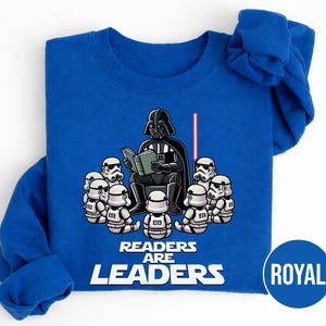 K&ouml;nnte beinhalten: K&ouml;nigsblaues Sweatshirt mit einer Grafik von Darth Vader, der Stormtroopern vorliest. Der Text "Readers are Leaders" ist unter der Illustration aufgedruckt. Ein rotes Lichtschwert ist ebenfalls zu sehen.