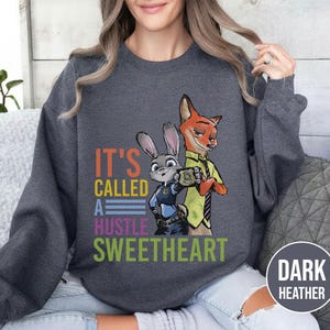 Puede incluir: Sudadera gris oscuro con un gr&aacute;fico colorido de los personajes de Zootopia, Judy Hopps y Nick Wilde. El texto "IT'S CALLED A HUSTLE SWEETHEART" est&aacute; impreso debajo de los personajes. La sudadera es de cuello redondo.