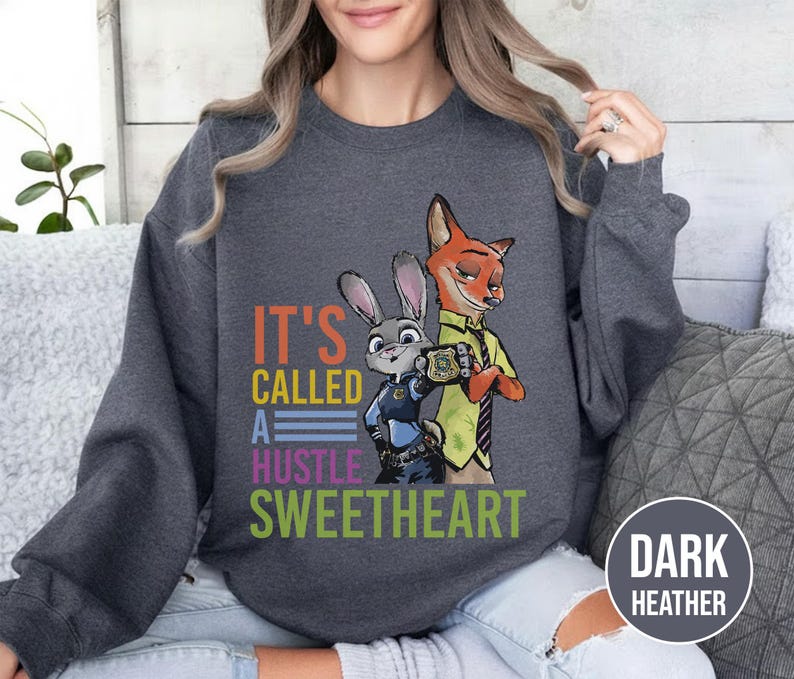 Puede incluir: Sudadera gris oscuro con un gr&aacute;fico colorido de los personajes de Zootopia, Judy Hopps y Nick Wilde. El texto "IT'S CALLED A HUSTLE SWEETHEART" est&aacute; impreso debajo de los personajes. La sudadera es de cuello redondo.