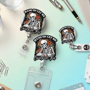 May include: Three badge reels featuring a skeleton illustration with orange and pink flowers, a book, and a steaming cup. The text reads "HANG ON LET ME Overthink This" in white lettering against a black glittery background with a clear plastic holder.