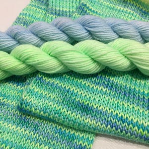 Puede incluir: Dos ovillos de hilo en tonos azules y verdes, y una muestra de tejido de punto en un patrón verde y azul jaspeado.