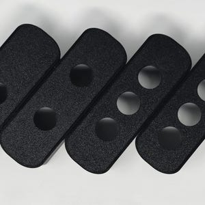 Könnte beinhalten: Vier schwarze, rechteckige Objekte mit abgerundeten Enden, die jeweils kreisförmige Aussparungen aufweisen. Die Objekte sind diagonal angeordnet, wobei die Anzahl der Aussparungen von eins bis drei zunimmt. Die Oberfläche hat ein strukturiertes Aussehen.