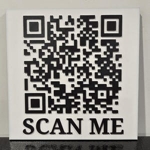 Könnte beinhalten: Ein quadratisches, weißes Schild mit einem großen schwarzen QR-Code-Design. Die Worte "SCAN ME" sind unten in Schwarz gedruckt. Der QR-Code besteht aus schwarzen Quadraten und Rechtecken und erzeugt ein einzigartiges Muster.