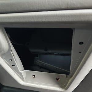 Op de afbeelding: Een rechthoekig, open compartiment verzonken in een lichtgrijs voertuigdashboard. Het compartiment is leeg, met zichtbare schroefgaten. De binnenkant is donker. Het omliggende dashboard heeft een getextureerd oppervlak.