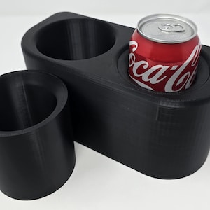 Könnte beinhalten: Ein schwarzer, 3D-gedruckter Auto-Konsolen-Organizer mit zwei Getränkehaltern und einem separaten zylindrischen Halter. Eine rote Coca-Cola-Dose befindet sich in einem der Getränkehalter. Der Organizer besteht aus schwarzem Kunststoff.