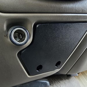 May include: Close-up of a black, textured panel with two circular holes, installed in a vehicle's interior. A silver-rimmed cigarette lighter receptacle is visible to the left. The panel is set against a gray, textured dashboard.