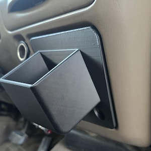 May include: A black, rectangular, 3D-printed storage bin, mounted on a black plate, attached to a vehicle's dashboard. The bin is square-shaped and open at the top. The dashboard is tan.