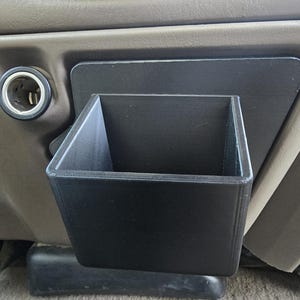 Op de afbeelding: Een zwarte, 3D-geprinte auto-console-organizer. De organizer is rechthoekig met een vierkante opening en wordt op het dashboard van een voertuig gemonteerd. Het ontwerp bevat een ingebouwde montageplaat voor eenvoudige installatie. De organizer is ontworpen om kleine items in op te bergen.