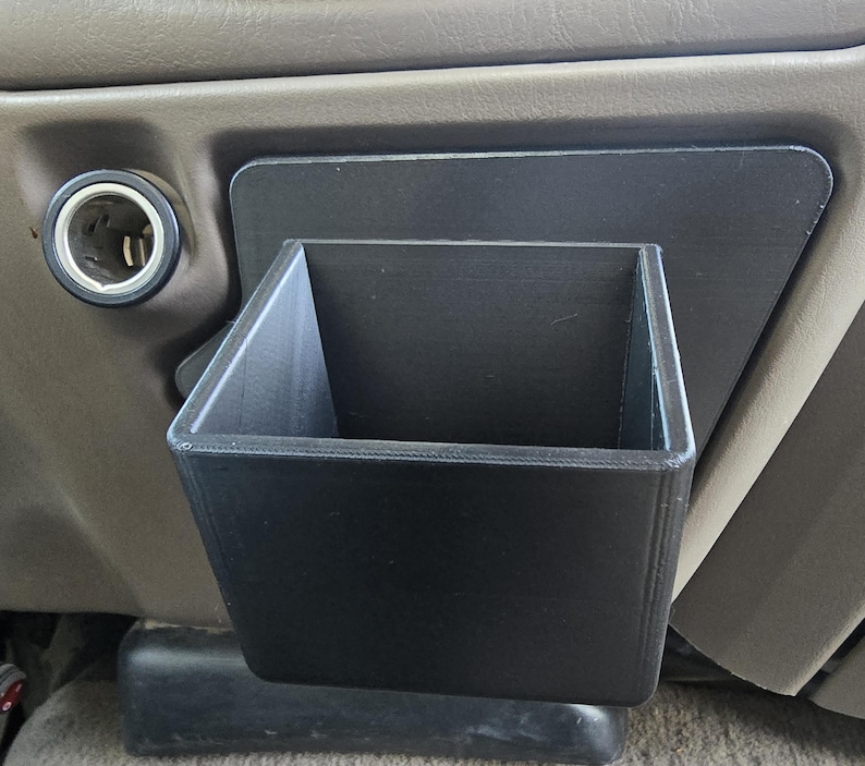 Op de afbeelding: Een zwarte, 3D-geprinte auto-console-organizer. De organizer is rechthoekig met een vierkante opening en wordt op het dashboard van een voertuig gemonteerd. Het ontwerp bevat een ingebouwde montageplaat voor eenvoudige installatie. De organizer is ontworpen om kleine items in op te bergen.