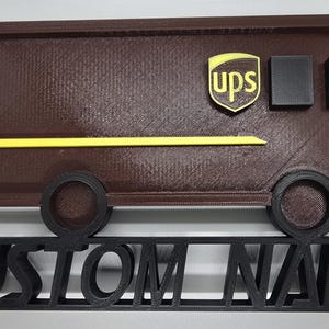Könnte beinhalten: Ein brauner 3D-gedruckter UPS-LKW mit gelbem Streifen und Logo sowie schwarzen Rädern. Darunter befindet sich ein schwarzes Schild mit der Aufschrift "CUSTOM NAME".
