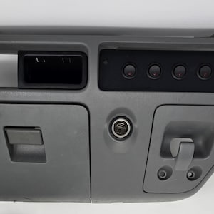 May include: A gray automotive dashboard center console panel featuring a rectangular storage compartment, a black switch panel with five circular buttons, a central circular power outlet socket, and a hinged storage door with a latch mechanism.