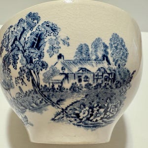 May include: Cream-colored ceramic bowl with a blue pastoral scene. The design features a house, trees, and a river, reminiscent of classic blue and white china. The bowl has a rounded shape and a slightly cracked surface.