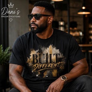 May include: A black short-sleeved t-shirt featuring a graphic print with the text "BUILT DIFFERENT" in a bold, distressed font. The design incorporates abstract gold, brown, and black paint splatters and brushstroke textures across the chest area.