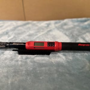 May include: A black and red digital torque wrench with the brand name "Snap-on" printed on the handle. The wrench has a digital display and a black grip. The tool is designed for precision tightening of fasteners.