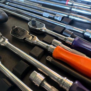 May include: A selection of chrome ratchets and extensions with black, orange, and purple handles. The tools are neatly arranged in a black case, displaying various sizes and styles. The image is well-lit, emphasizing the metallic finish of the tools.