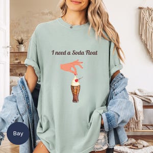 Könnte beinhalten: Ein hellblaues T-Shirt mit dem Text "I need a Soda Float" und einer Grafik einer Hand, die eine Kirsche auf einen Soda Float legt.