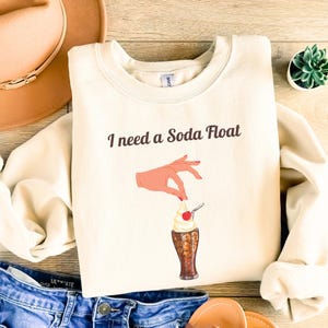 Könnte beinhalten: Ein cremefarbener Pullover mit dem Text "I need a Soda Float" und einer Grafik einer Hand, die eine Kirsche über einem Glas Soda mit Schlagsahne hält.