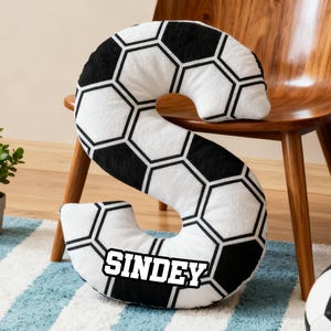 Puede incluir: Un cojín blanco y negro con temática de balón de fútbol en forma de la letra "S". El cojín tiene un patrón de balón de fútbol y el nombre "SINDEY" está impreso en él. El cojín está sobre una alfombra a rayas azules y blancas.