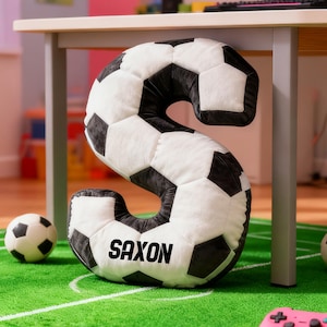 Puede incluir: Un gran cojín con forma de letra "S" con forma de balón de fútbol, con el nombre "SAXON" impreso. El cojín es blanco y negro, con un diseño de balón de fútbol. Dos balones de fútbol y un mando de juego rosa están sobre una alfombra verde.